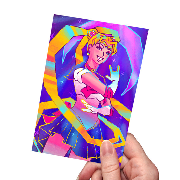 Hand holding a postcard-sized art print featuring a girl in a sailor outfit striking a pose with golden hair spiraling around her. stars fill the background and a large crescent moon hangs behind her. 